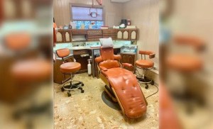 A wide shot of a retro-style dental treatment room featuring an orange upholstered patient chair and matching stools. The room is furnished with classic wood-grain cabinetry, a beige tiled floor, and traditional dental equipment, giving it a mid-century aesthetic.
